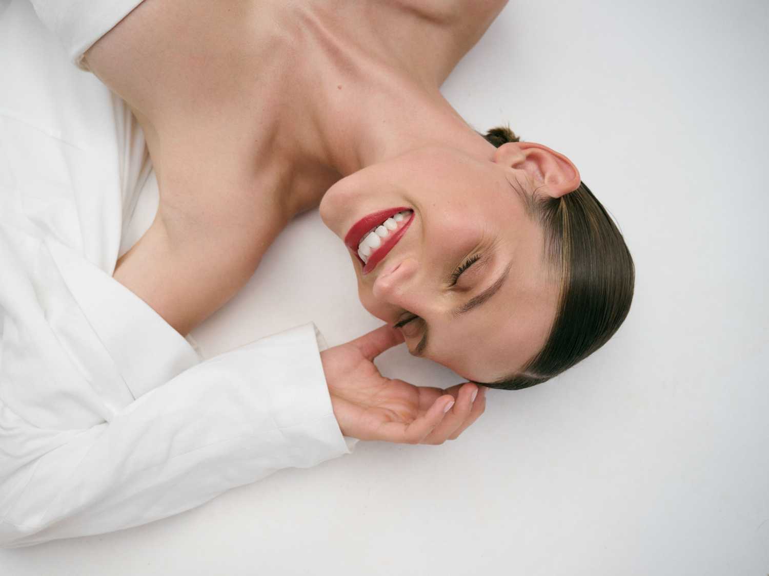 Na białym tle leży uśmiechnięta kobieta w białej koszuli z zamkniętymi oczami i czerwoną szminką na ustach, trzymająca dłoń przy twarzy.