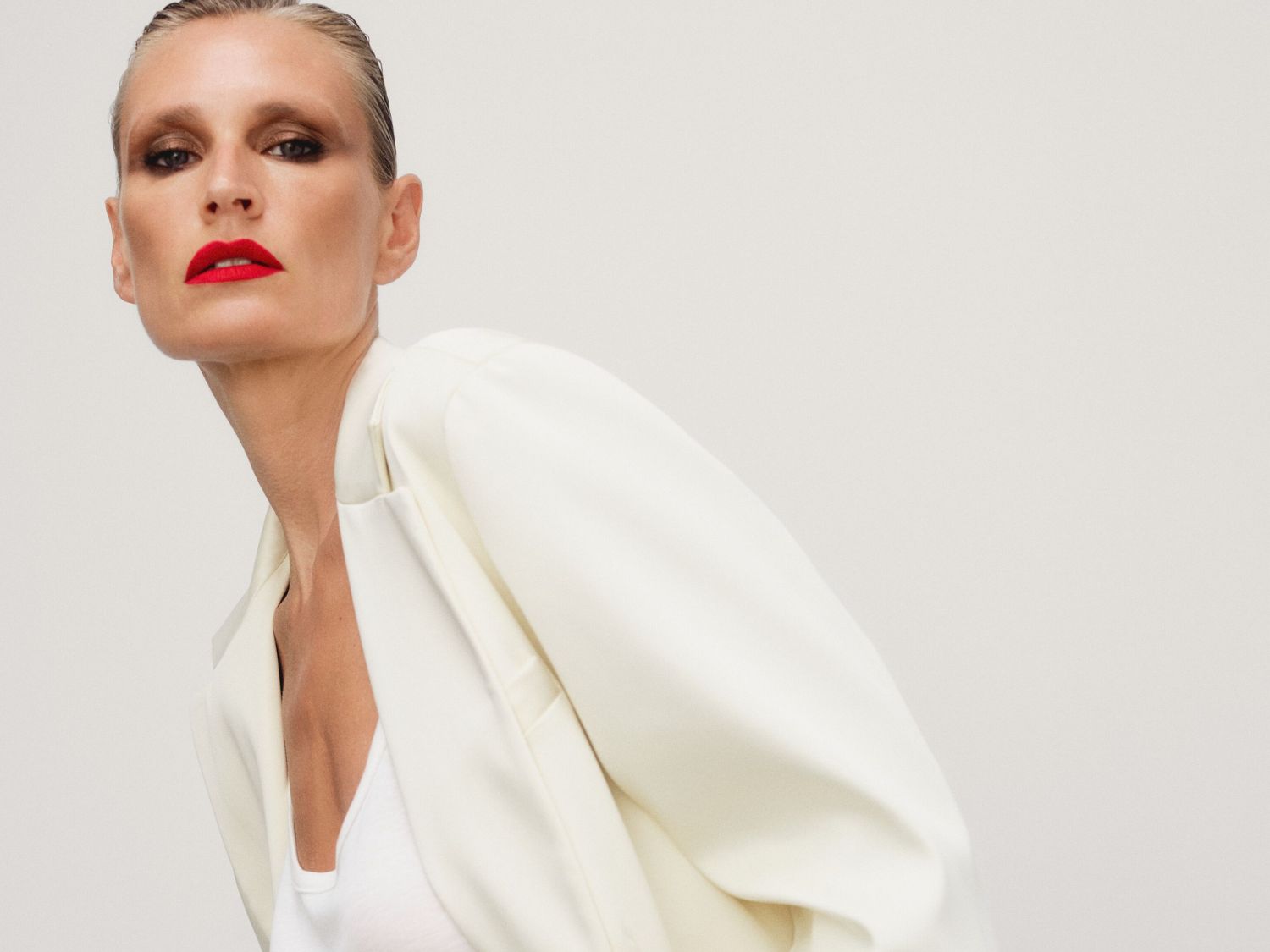 Portret kobiety z mokrymi włosami, w białym topie i marynarce, z czerwoną szminką na ustach, pozującej na jasnym tle.