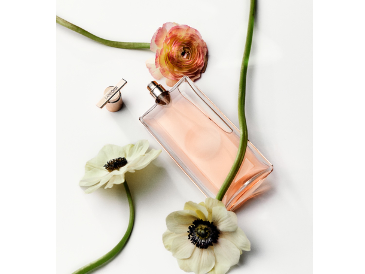 Perfumy Lancôme Idôle w przezroczystej butelce z różową zawartością, otoczone białymi i różowymi kwiatami na białym tle.