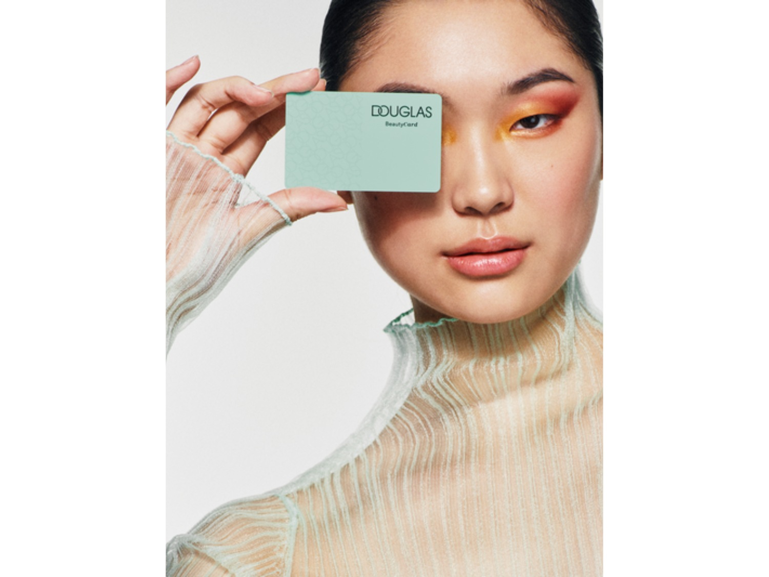 Portret kobiety z makijażem oczu w odcieniach pomarańczy i różu, trzymającej kartę Douglas Beauty przed okiem.