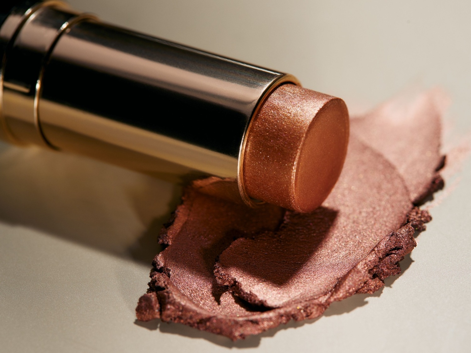 Sztyft do makijażu w złotym opakowaniu obok próbki produktu o metalicznym, brązowym odcieniu, ukazujący jego kremową konsystencję i połysk.