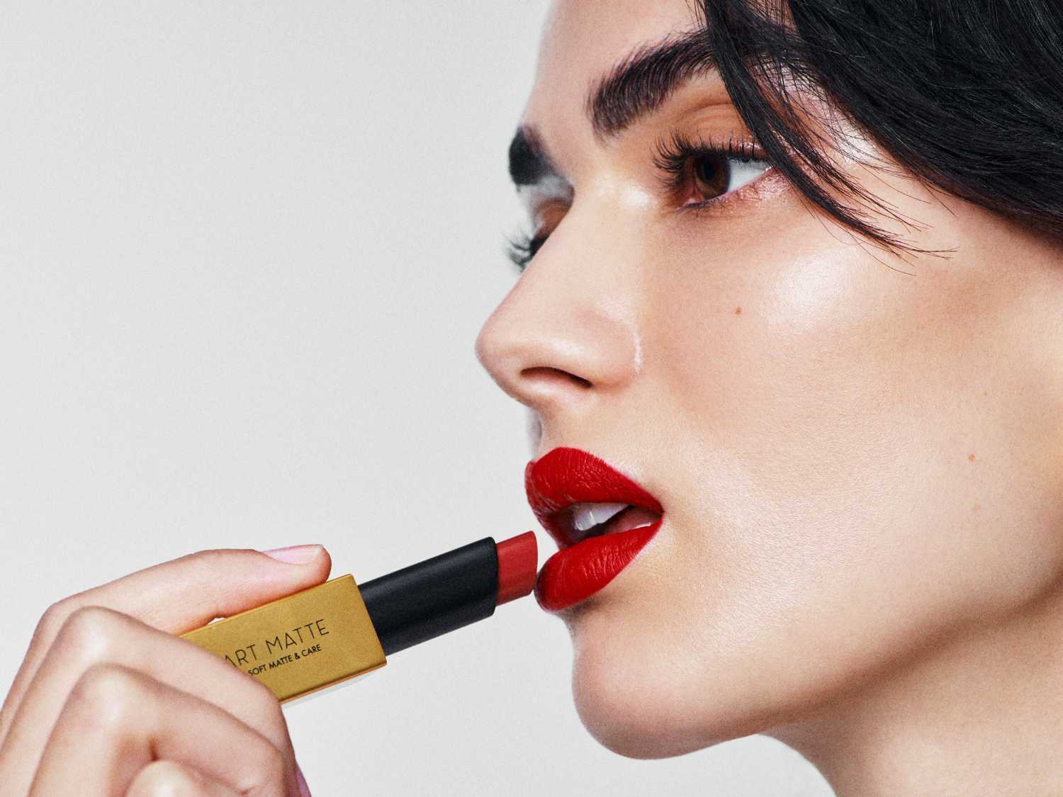 Kobieta z ciemnymi włosami aplikuje czerwoną szminkę Art Matte z opakowania w kolorze złotym i czarnym, aby uzyskać miękkie, matowe wykończenie.