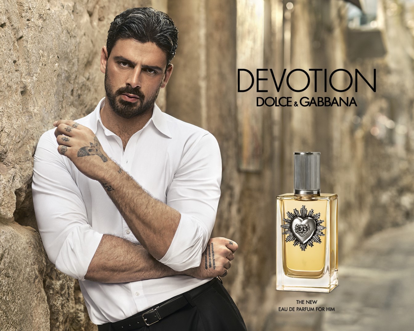 A Dolce & Gabbana Devotion Eau de Parfum for Him reklámképe: egy sármos férfi fehér ingben egy kőfalnak támaszkodik, mellette az aranyszínű parfümös üveg ezüst szívvel.