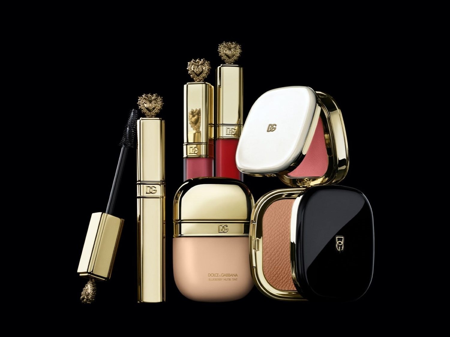 A Dolce & Gabbana sminktermékek kollekciója, köztük arany szempillaspirál, rúzs, alapozó és púder, elegáns csomagolásban, fekete háttér előtt.