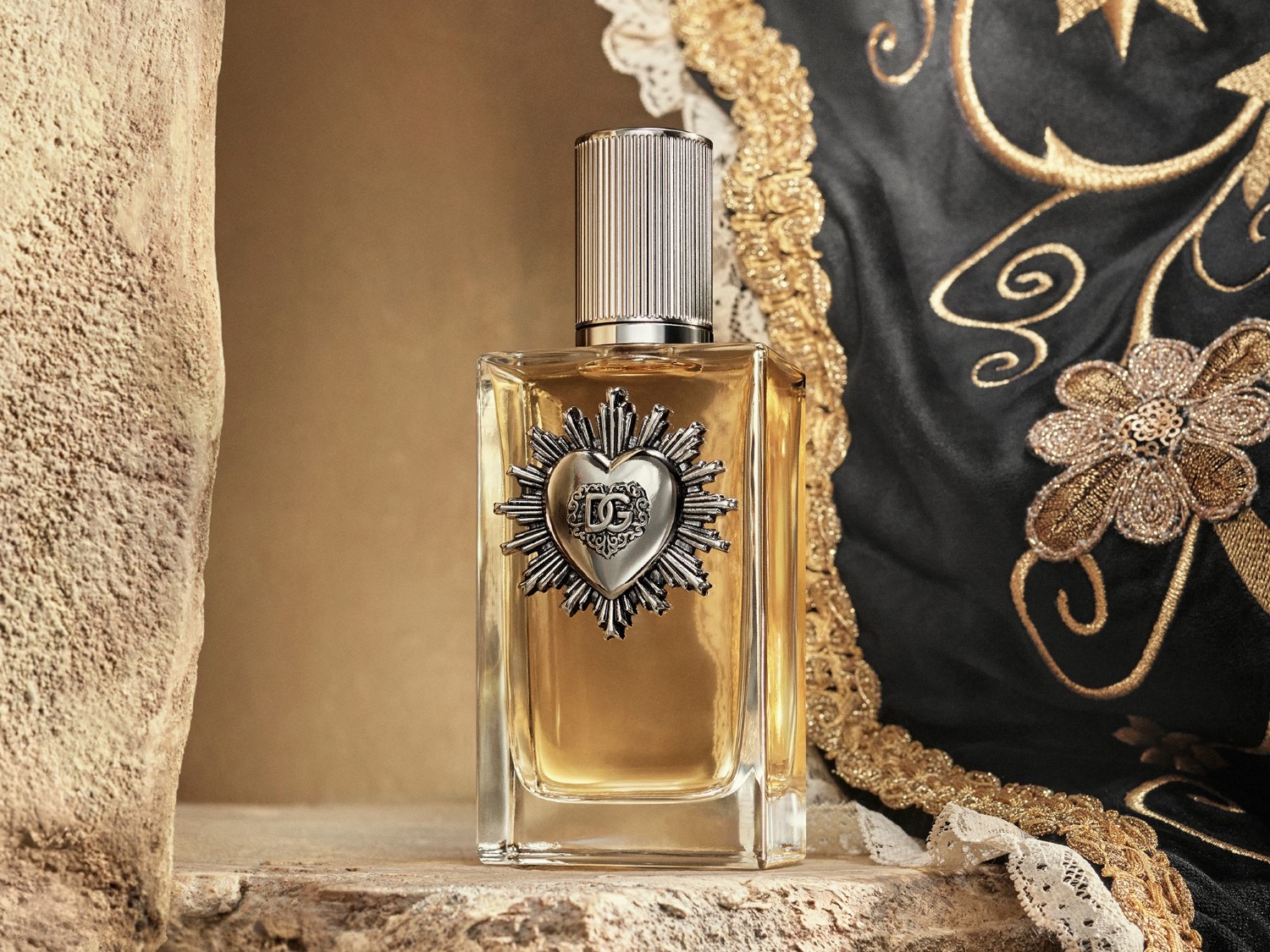 A Dolce & Gabbana Devotion Eau de Parfum üvegben, aranyszínű folyadékkal, ezüst szív alakú logóval, texturált ezüst kupakkal, fekete és arany hímzett anyag mellett.