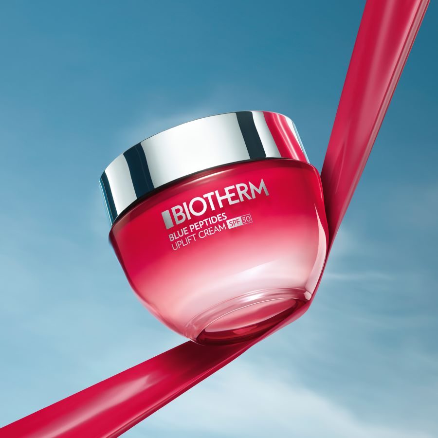 Biotherm Blue Therapy Uplift Creme mit LSF 30 in einem transparenten, rot getönten Tiegel mit silbernem Deckel, der auf einem roten Band vor blauem Himmel balanciert.