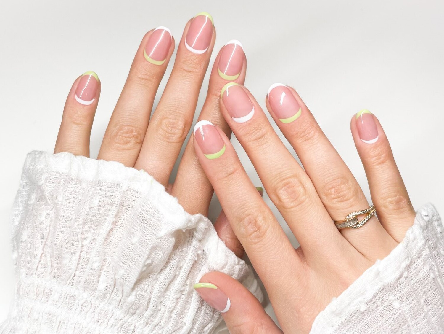 Nahaufnahme von Händen mit French Manicure, wobei die Nagelspitzen in Weiß und Limettengrün gehalten sind, ergänzt durch einen goldenen Ring.