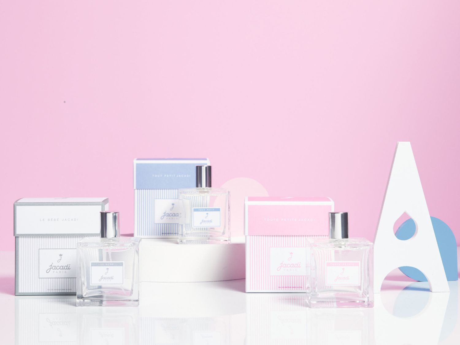 Na růžovém pozadí jsou zobrazeny tři flakony dětských parfémů Jacadi s bílými krabičkami s modrými a růžovými pruhy.