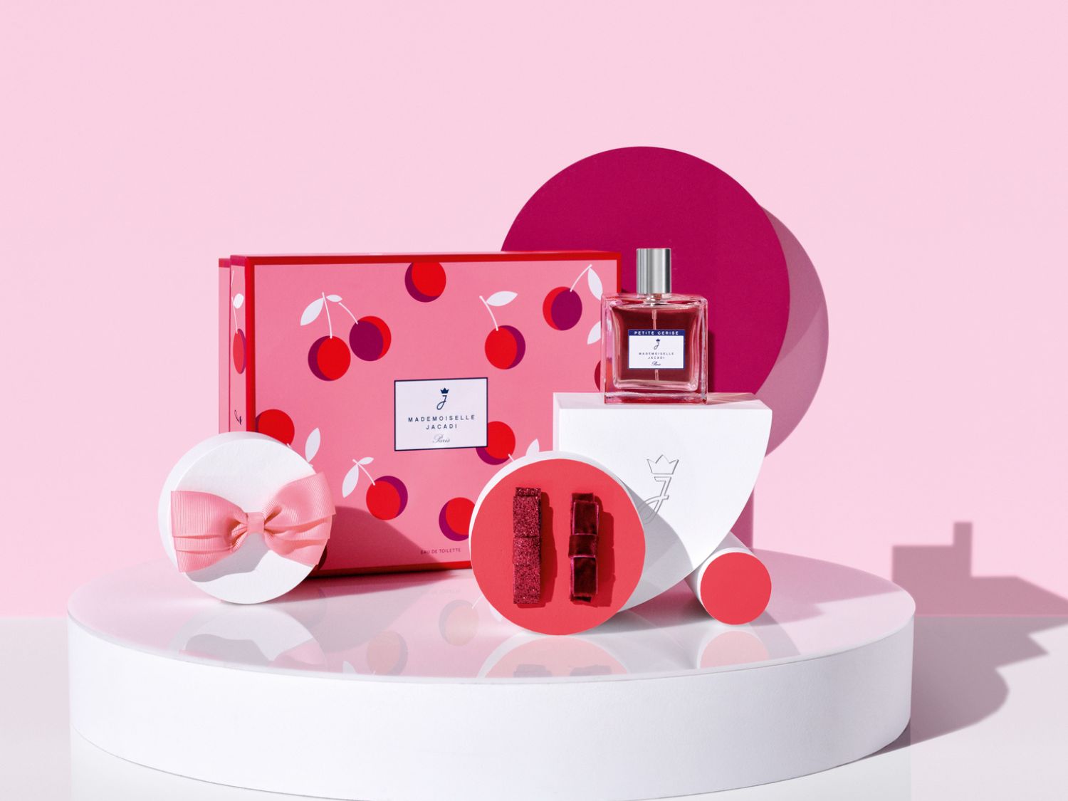 Růžový dárkový set Mademoiselle Jacadi Paris s třešňovým motivem, flakon parfému Petite Cerise a ozdobná mašle, vše na bílém podstavci.