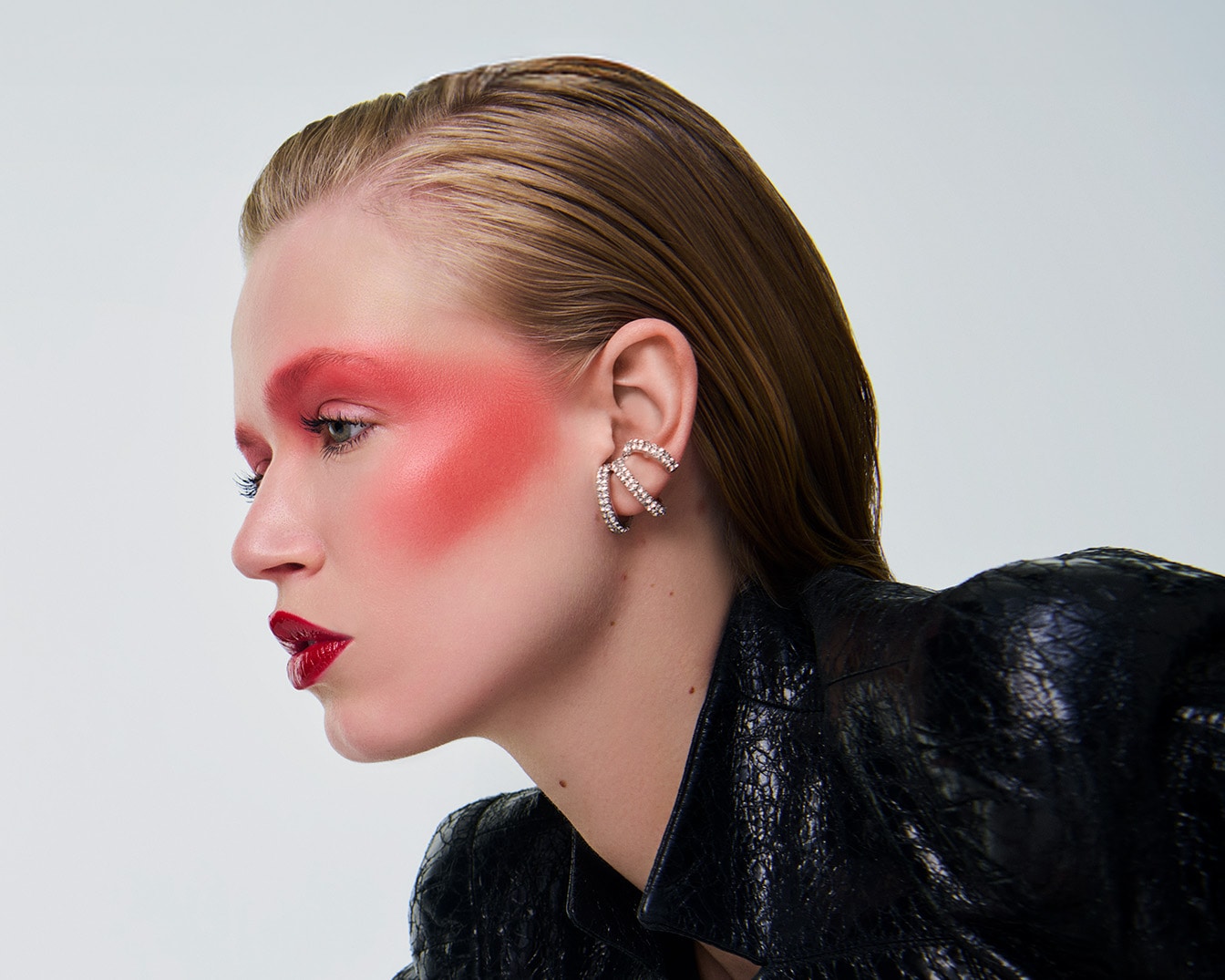 Une femme au maquillage audacieux, avec un fard à joues rouge vif s'étendant jusqu'aux tempes, des lèvres rouges brillantes et une veste en cuir noir, porte une boucle d'oreille en forme de C ornée de strass.