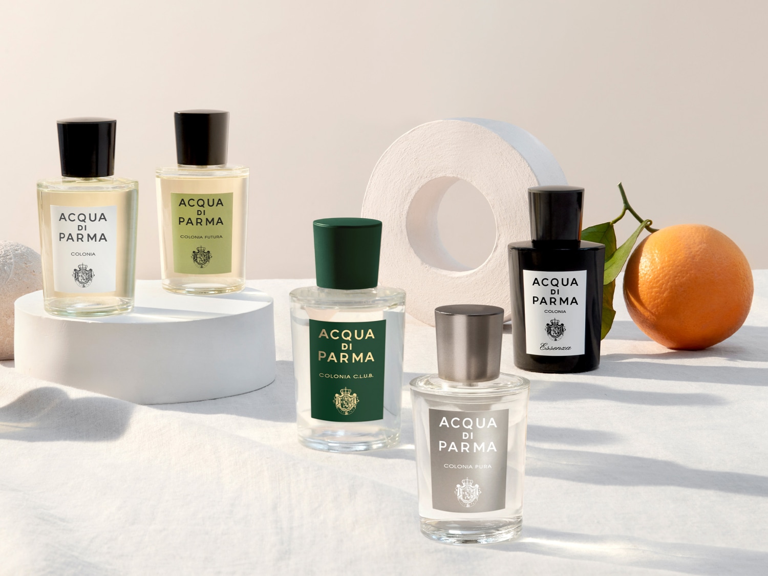 Pět lahví kolínské vody Acqua di Parma různých vůní, včetně Colonia, Colonia Futura, Colonia Club, Colonia Pura a Essenza, uspořádaných s pomerančem a bílým prstenem.