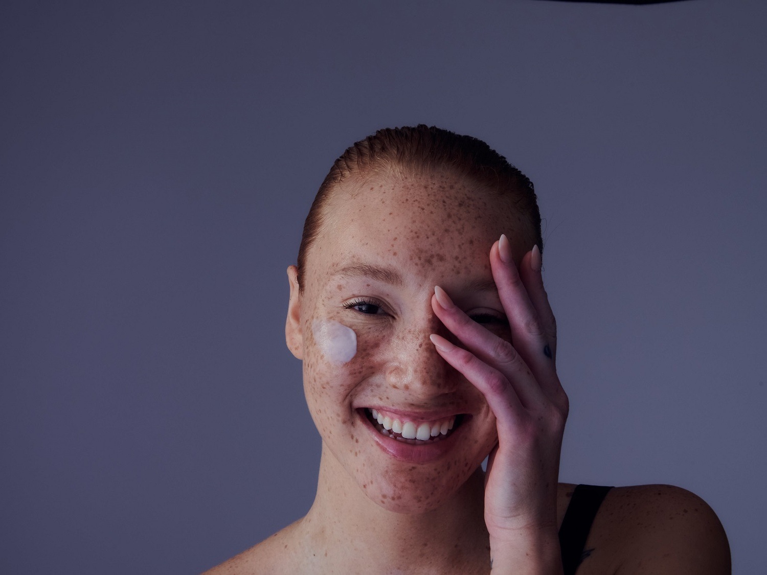 Une femme rousse souriante, avec une touche de crème blanche sur la joue, cache une partie de son visage avec sa main, sur un fond gris.