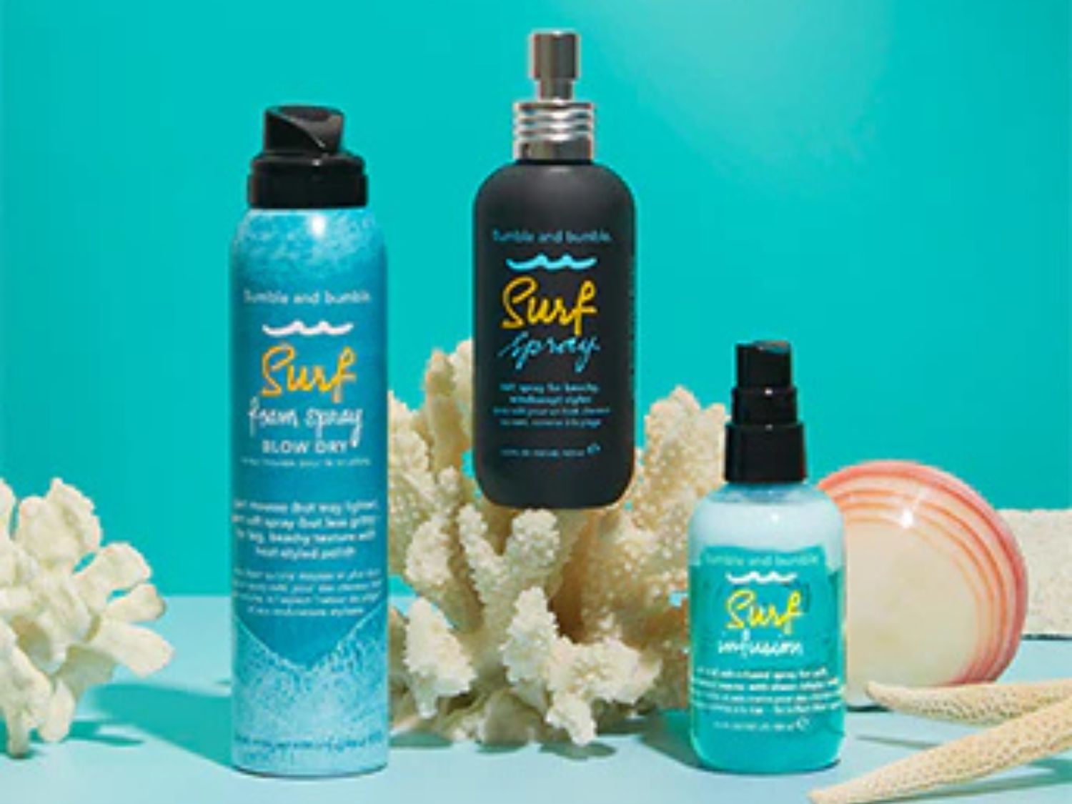 Tři produkty péče o vlasy Bumble and Bumble Surf, včetně spreje, pěny a infuze, na pozadí korálů a mořských mušlí.
