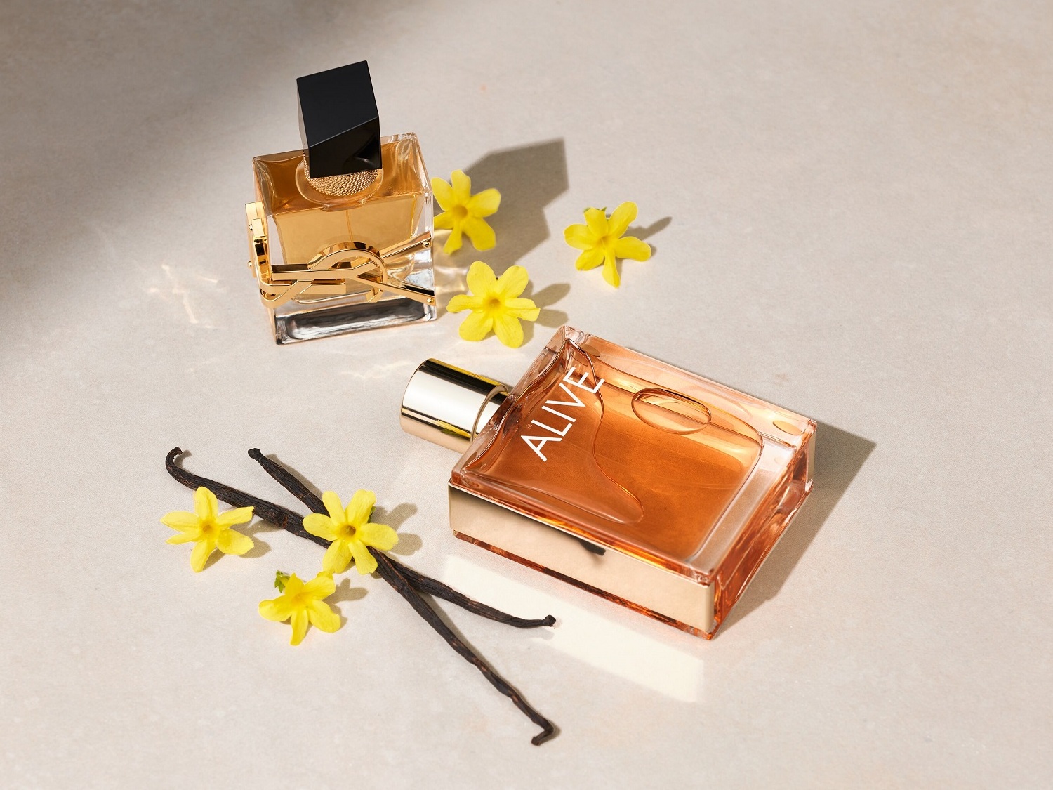 Dva flakóny parfumov, jeden Yves Saint Laurent s čiernym vrchnákom a druhý Hugo Boss Alive, sú obklopené žltými kvetmi a vanilkovými strukmi.