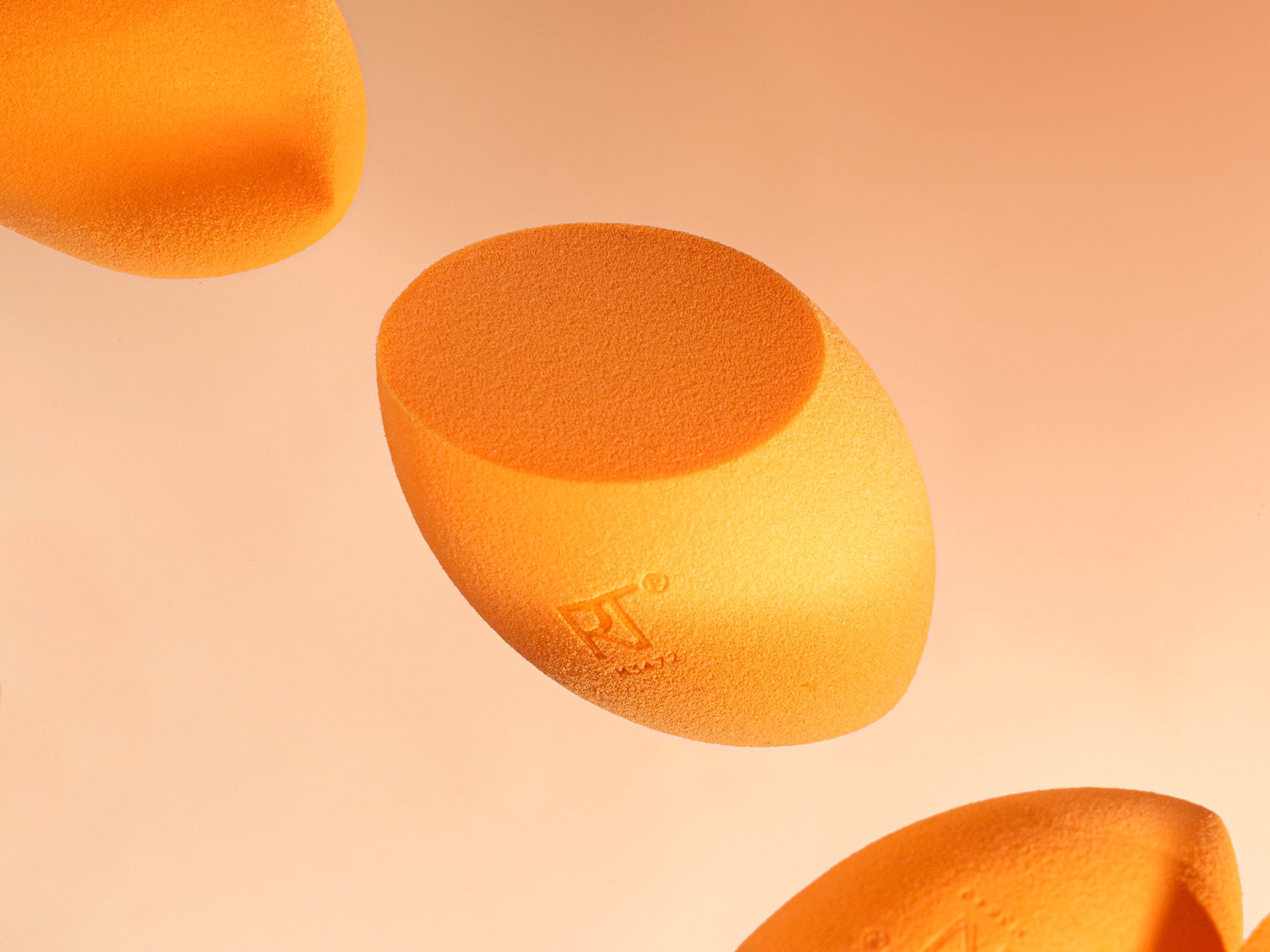 Nahaufnahme von orangefarbenen Make-up-Schwämmchen mit dem RT-Logo, die auf einem hellorangefarbenen Hintergrund schweben.