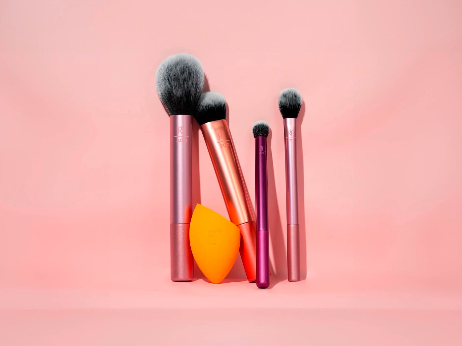 Auf einem rosa Hintergrund sind vier Make-up-Pinsel mit rosa, kupferfarbenen und violetten Griffen sowie ein orangefarbener Make-up-Schwamm zu sehen.