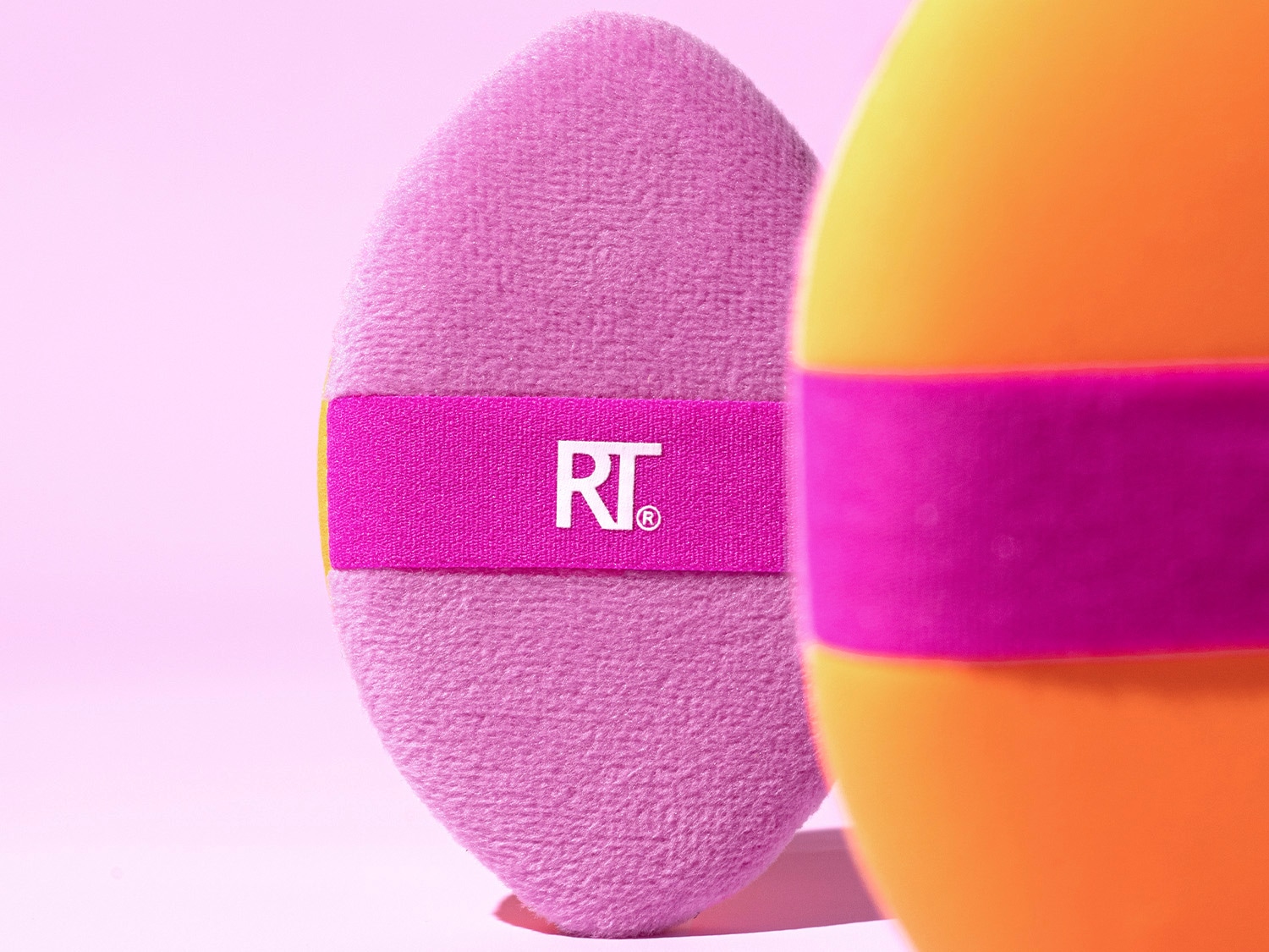 Nahaufnahme eines rosa Make-up-Pads mit einem pinkfarbenen Band mit dem RT-Logo, daneben ein orangefarbener Make-up-Schwamm mit einem pinkfarbenen Band.