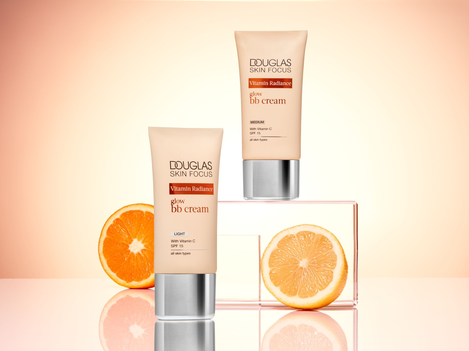 Dwa odcienie kremu BB Douglas Skin Focus Vitamin Radiance Glow: Light i Medium, oba z witaminą C i SPF 15, obok nich połówki pomarańczy.