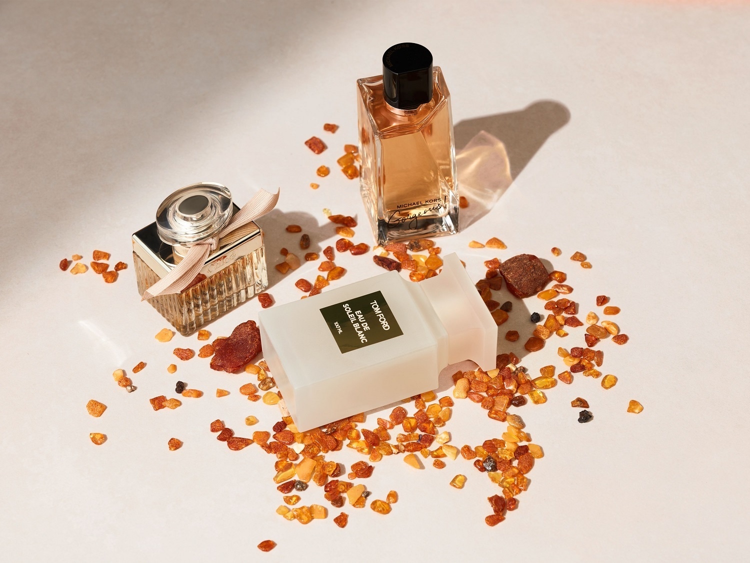 Tri parfumy od rôznych značiek, vrátane Chloe, Tom Ford a Michael Kors, sú usporiadané na bielom pozadí s rozhádzanými kúskami jantáru.