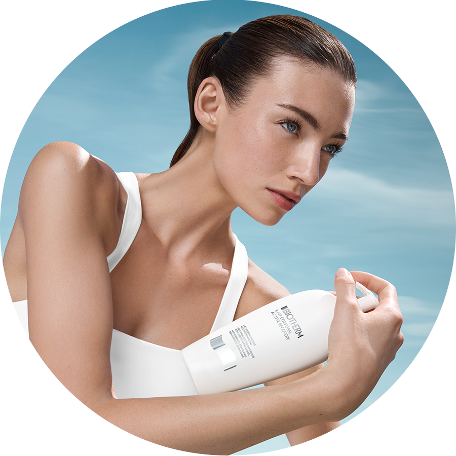 Eine Frau im weißen Sport-BH hält eine weiße Flasche Biotherm Lait Corporel Active Recovery vor einem hellblauen Himmel.