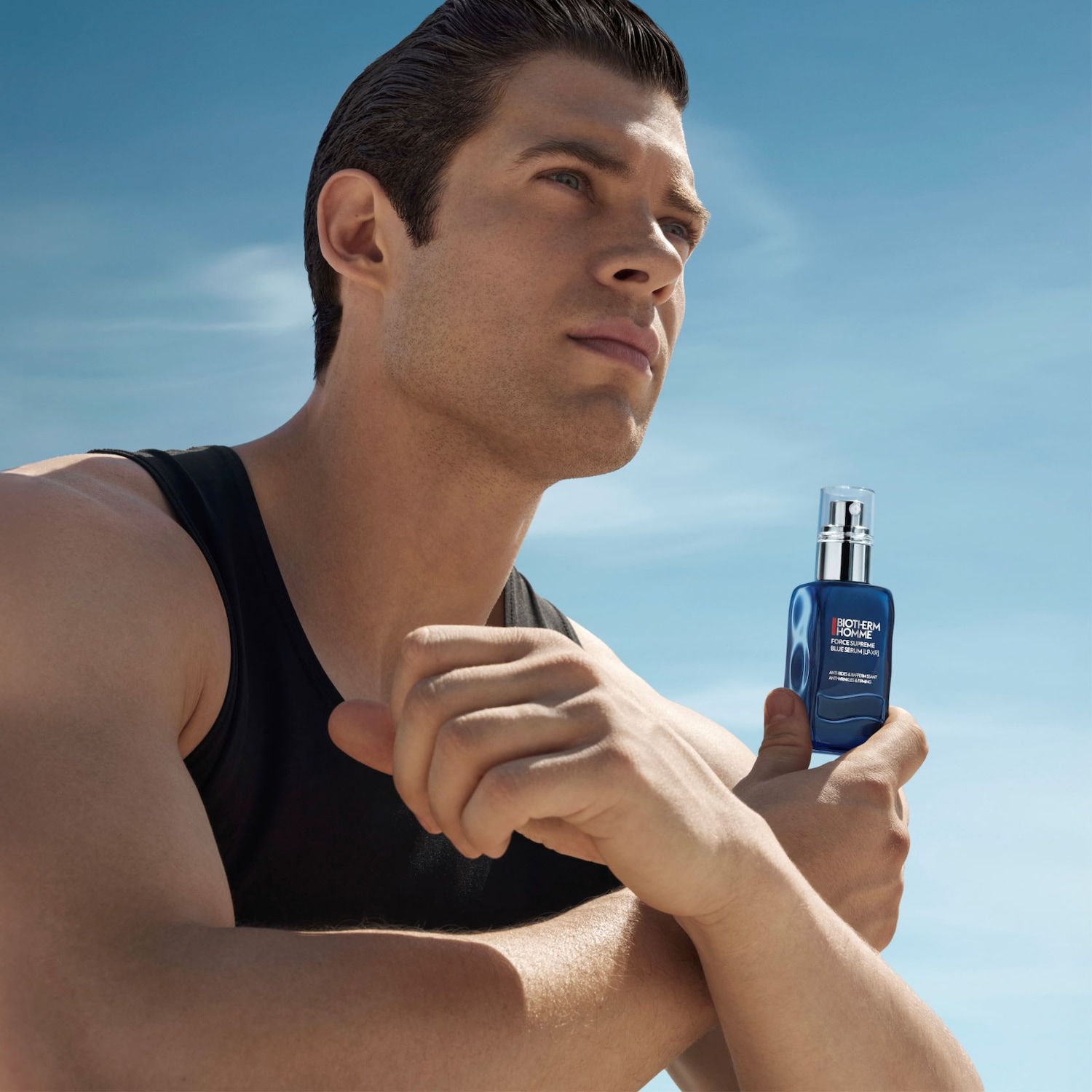 Ein Mann mit dunklem Haar und schwarzem Tanktop hält eine blaue Flasche Biotherm Homme Force Supreme Blue Serum in der Hand.