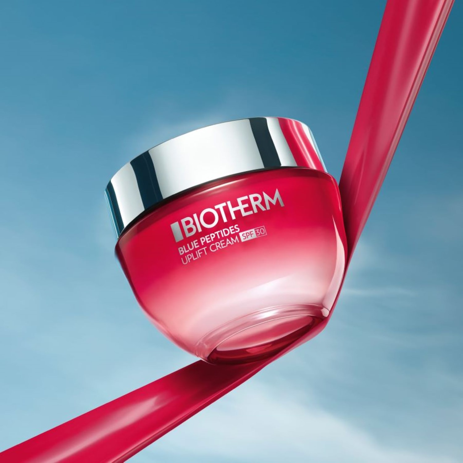 Nahaufnahme von Biotherm Blue Peptides Uplift Cream SPF 30 in einem rot-weißen Tiegel mit silbernem Deckel, der auf einem roten Band vor einem hellblauen Himmel balanciert.