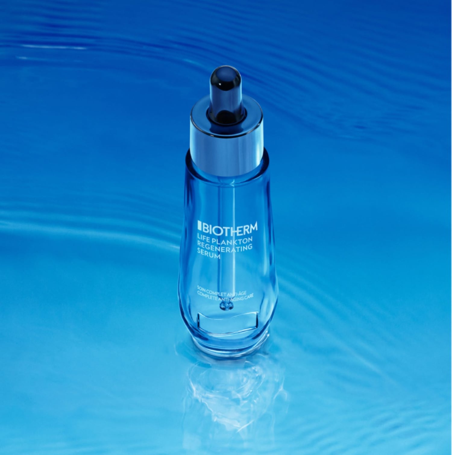 Das Biotherm Life Plankton Regenerating Serum in einer durchsichtigen, blauen Glasflasche mit Pipette, die auf einer glatten, blauen Oberfläche steht.