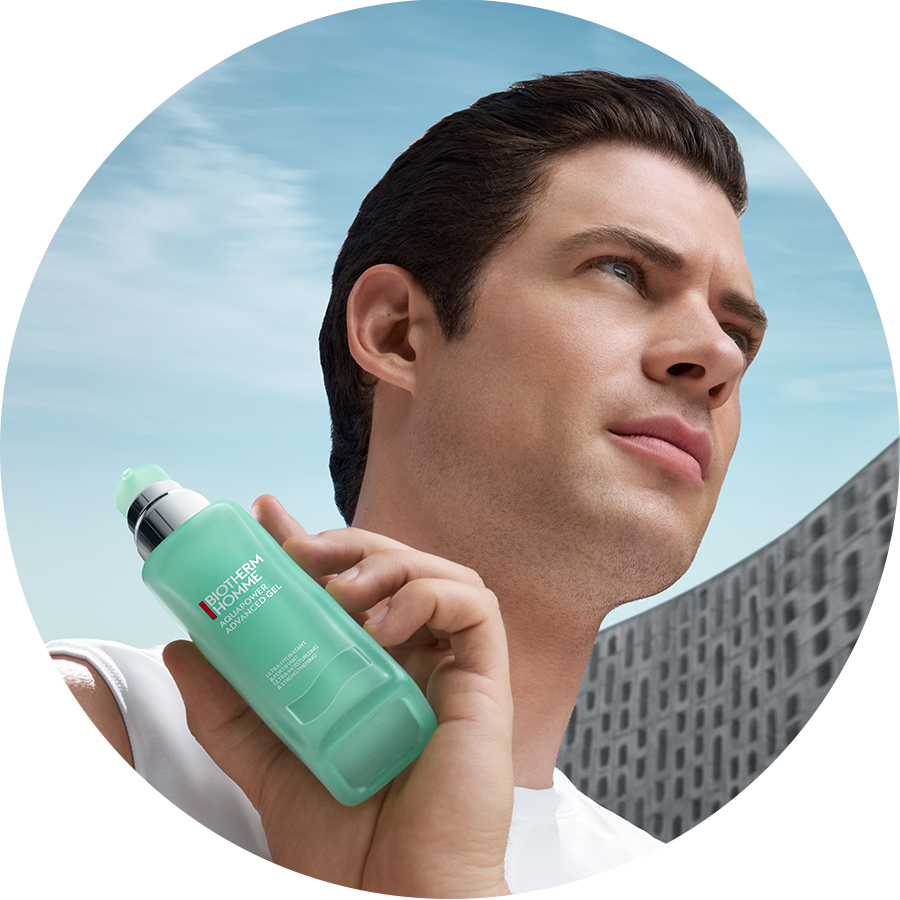 Ein Mann hält Biotherm Homme Aquapower Advanced Gel in einer hellgrünen Flasche mit silbernem Spender vor einem Hintergrund aus blauem Himmel und einem Gebäude.