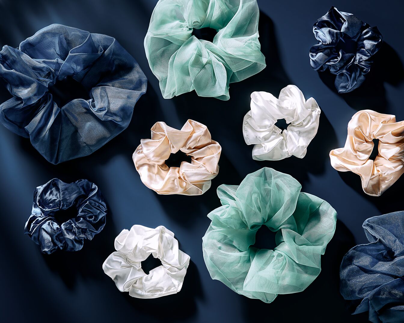 Une collection d'élastiques à cheveux en tissu soyeux bleu marine, vert d'eau, blanc et pêche, disposés sur un fond bleu foncé, offrant une touche d'élégance à votre coiffure.