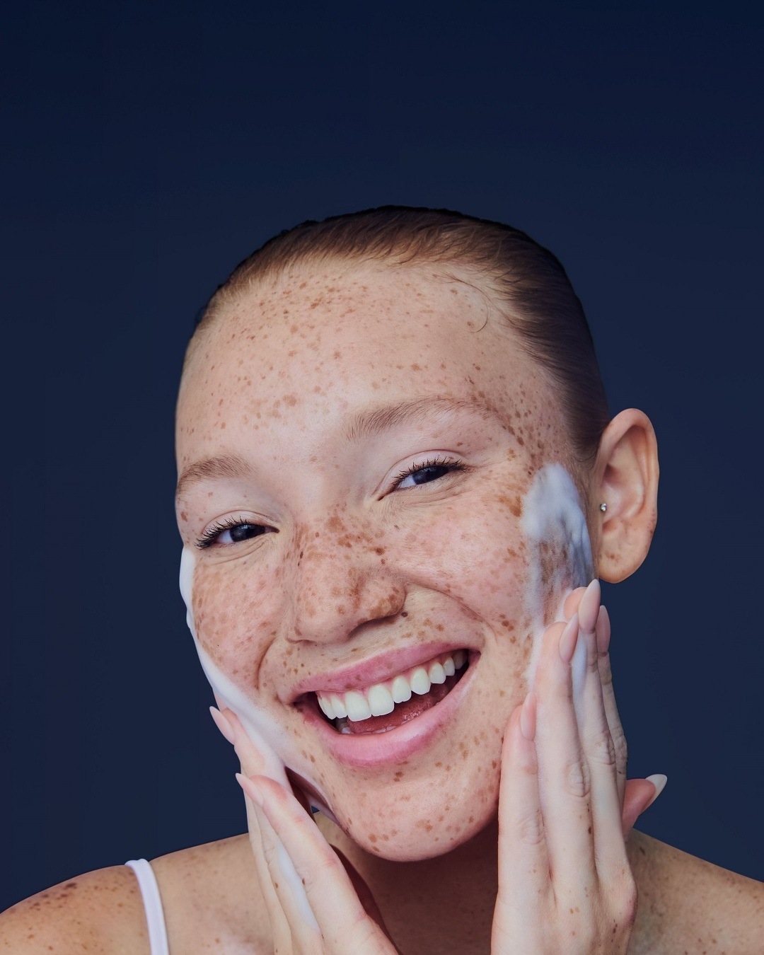 Une femme souriante, le visage couvert de mousse nettoyante, se masse doucement la peau pour un teint éclatant.