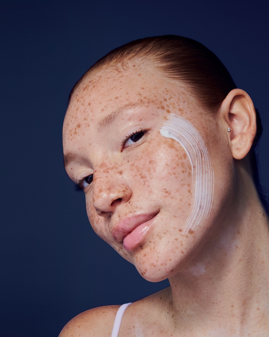 Une femme souriante applique une crème blanche sur sa joue, mettant en valeur sa peau naturellement éclatante.