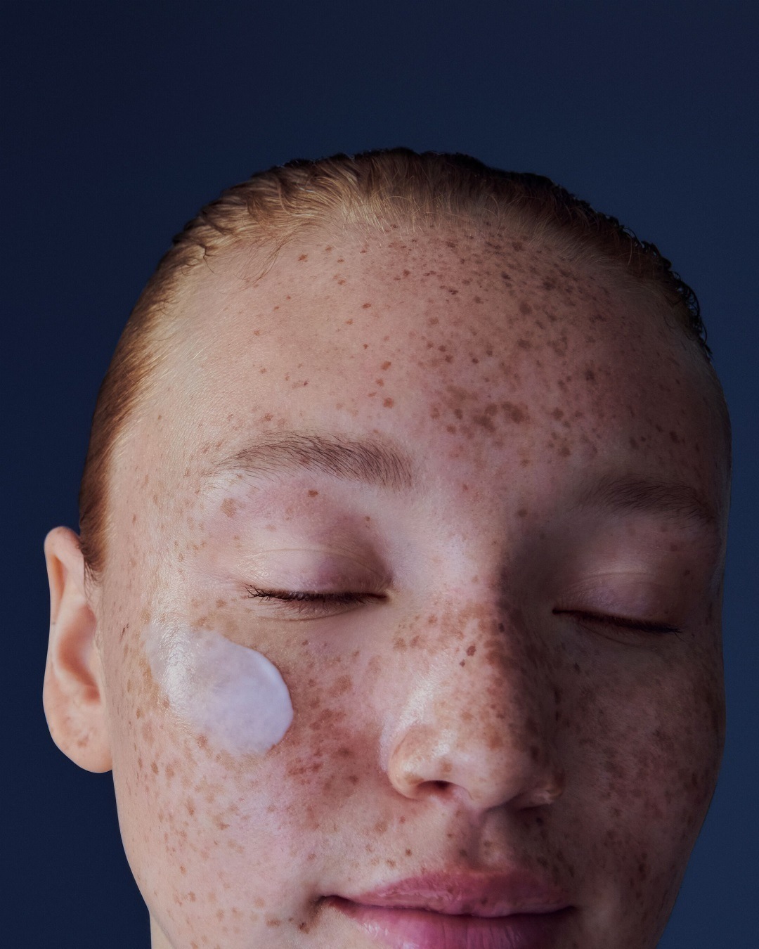 Une femme aux cheveux roux et à la peau parsemée de taches de rousseur, les yeux clos, avec une touche de crème blanche sur la joue.