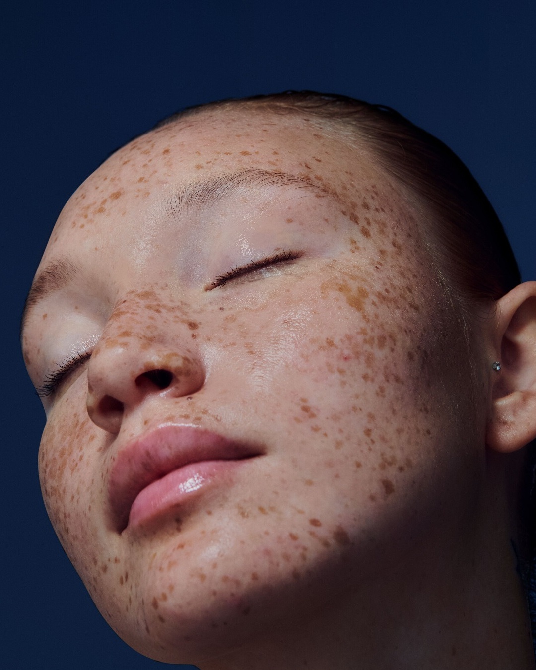 Gros plan sur le visage d'une jeune femme à la peau claire parsemée de taches de rousseur, les yeux clos et les lèvres brillantes, sur un fond bleu foncé.