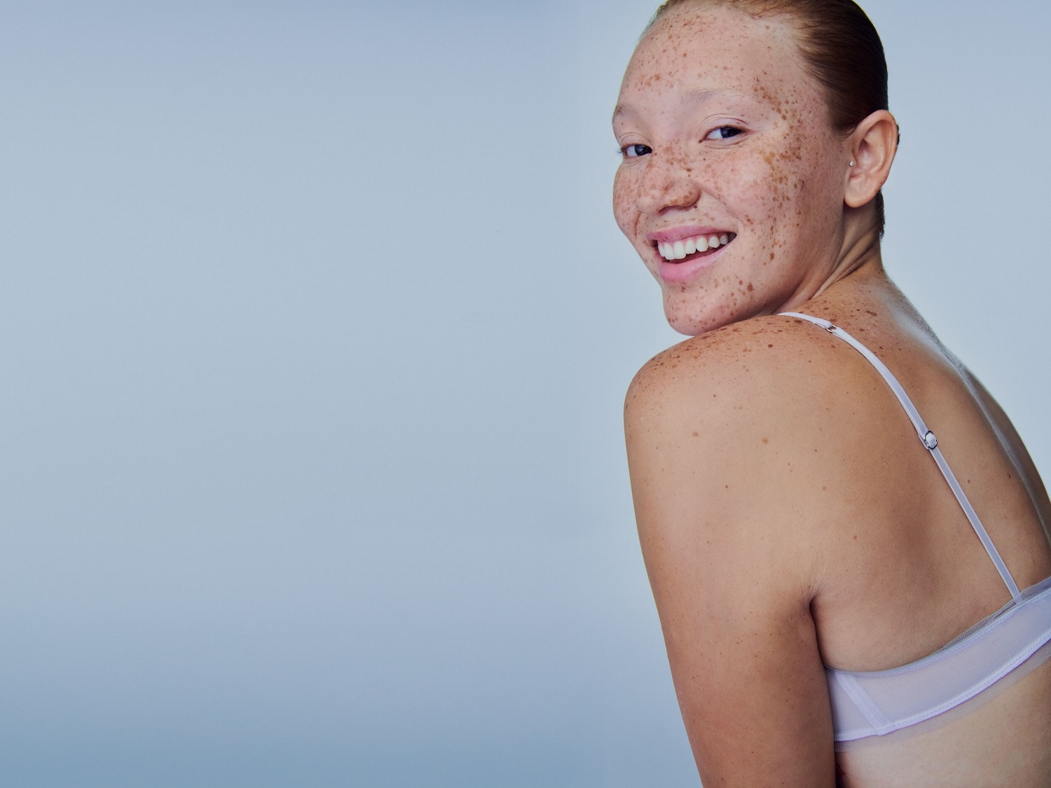 Une femme souriante aux cheveux roux et aux taches de rousseur, portant un soutien-gorge à fines bretelles, regarde par-dessus son épaule avec un sourire éclatant.