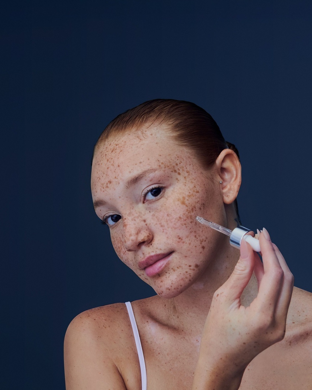 Une femme applique un sérum avec un compte-gouttes sur son visage, mettant en valeur une peau éclatante et des taches de rousseur naturelles.