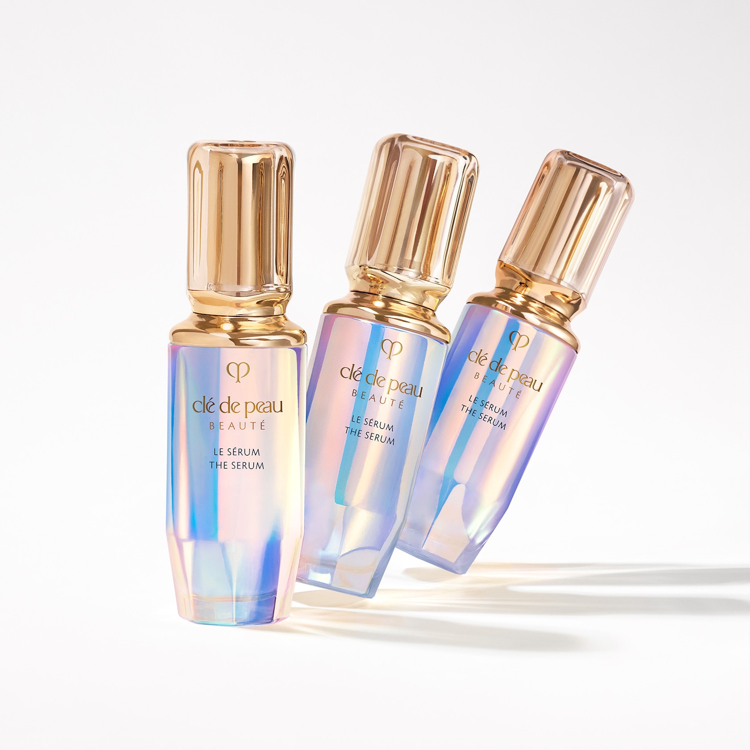 Tres frascos de suero Clé de Peau Beauté, con envases iridiscentes y tapas doradas, sobre un fondo blanco.