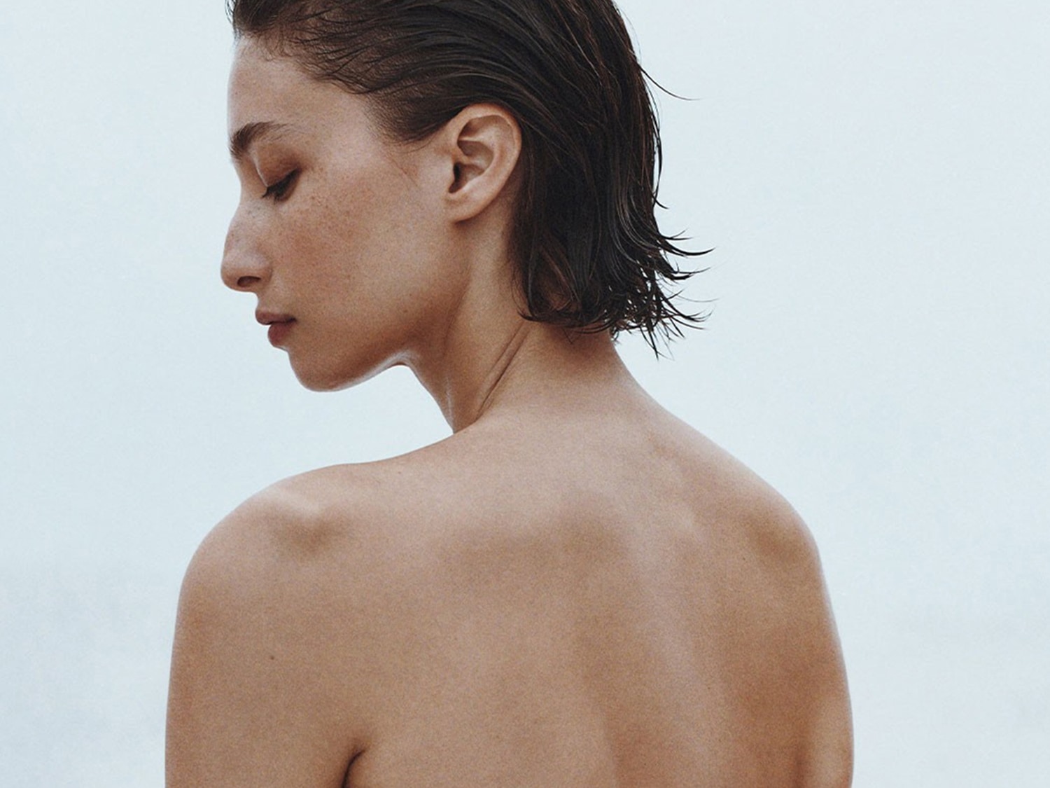 Na jasnym tle widać kobietę z krótkimi, mokrymi, ciemnymi włosami, odwróconą tyłem, z zamkniętymi oczami i delikatnym makijażem.