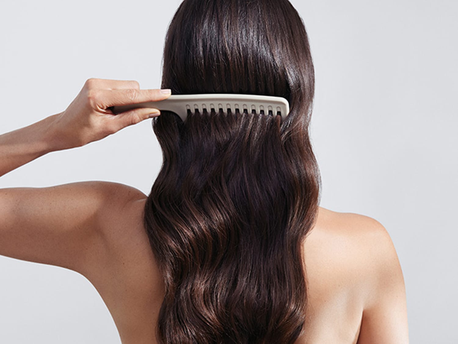Kobieta czesze swoje długie, brązowe włosy beżowym grzebieniem o szerokich zębach, stojąc tyłem do widza.
