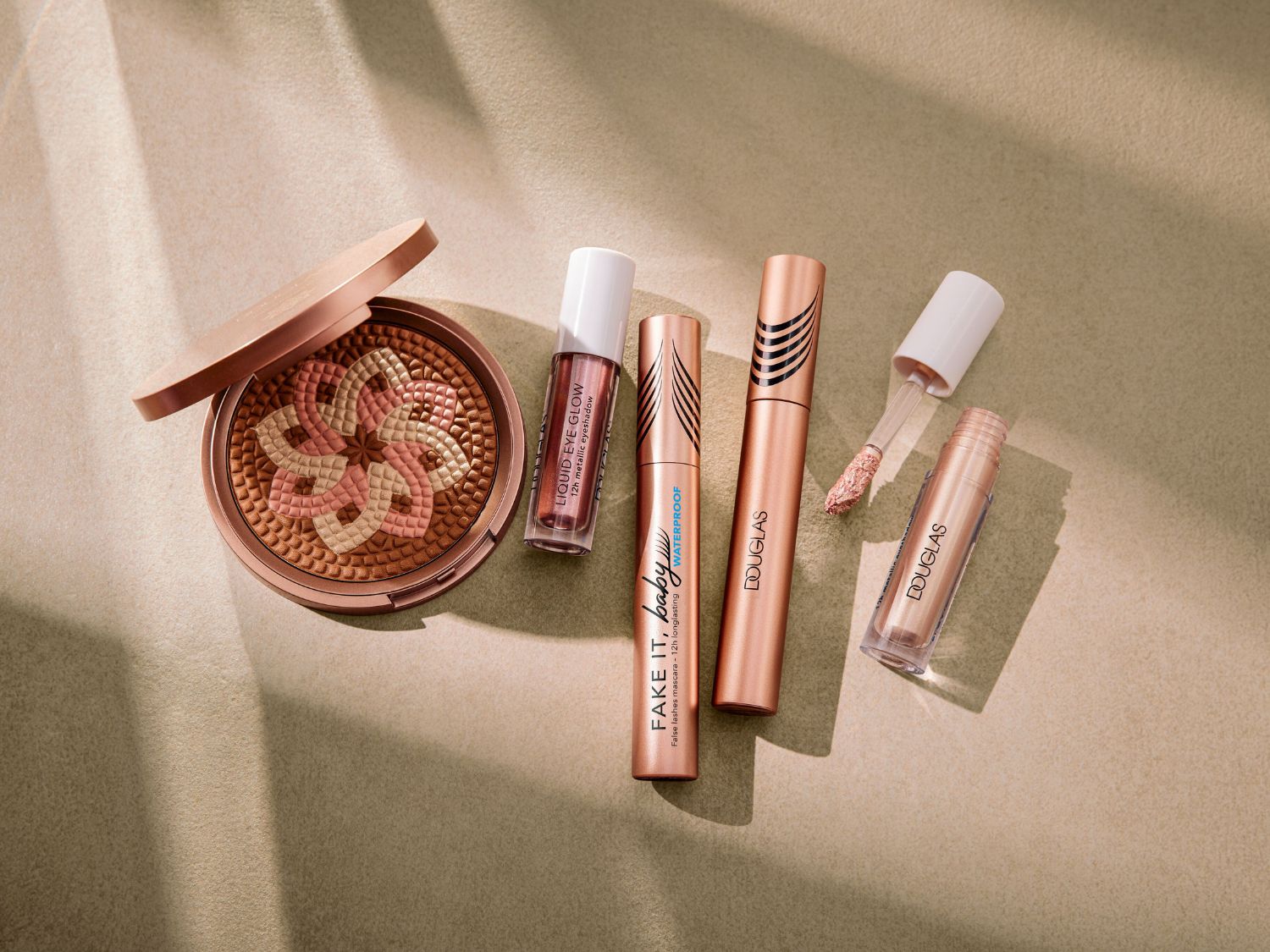 Kolekcja kosmetyków Douglas w odcieniach różu i brązu, w tym rozświetlacz do twarzy, tusz do rzęs Fake It Baby i metaliczny cień do powiek Liquid Eye Glow.