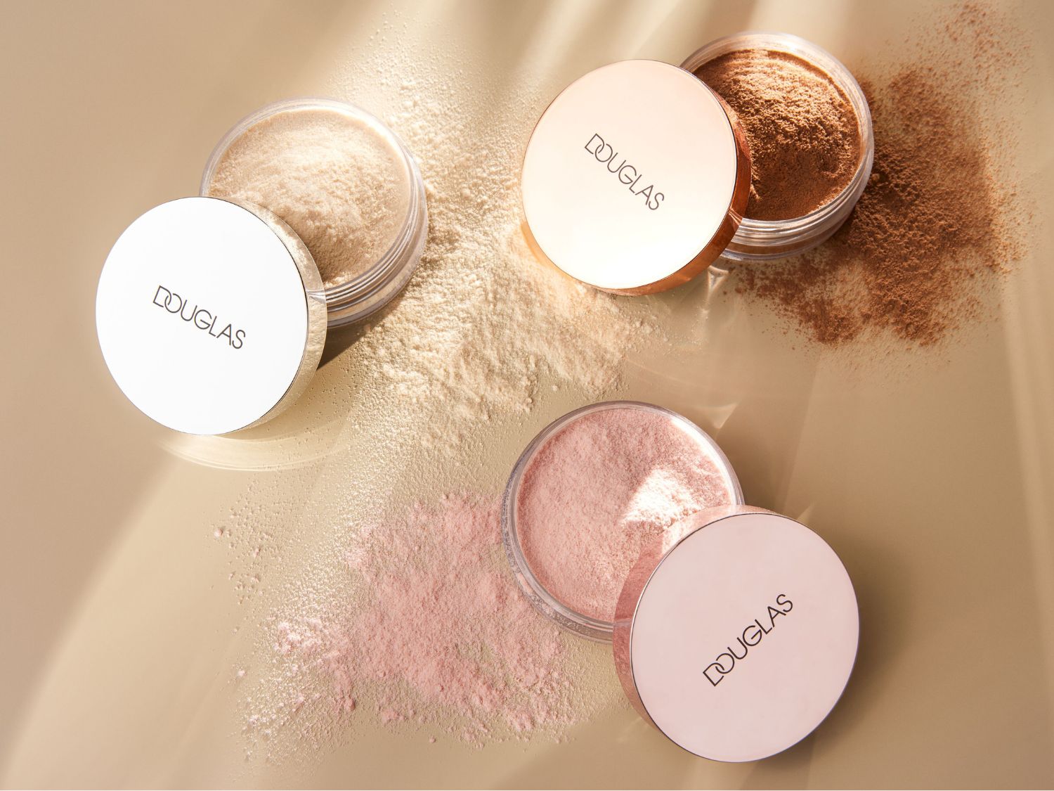 Pudry Douglas w odcieniach beżu, brązu i różu, każdy w przezroczystym słoiczku z wieczkiem w kolorze różowego złota, rozsypane na beżowym tle.