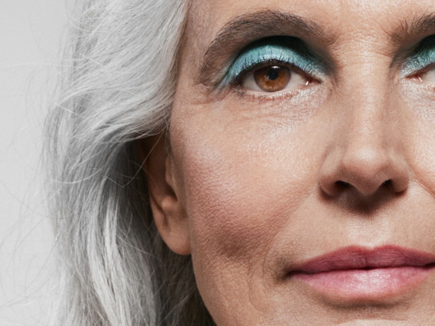 Nahaufnahme eines Frauengesichts mit grauen Haaren, braunen Augen und blauem Lidschatten, die Lippen sind mit einem rosafarbenen Lippenstift geschminkt.