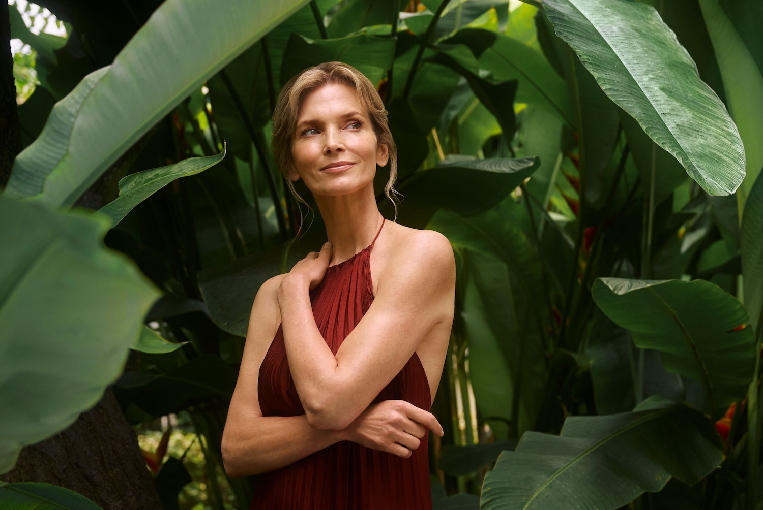 Eine Frau mit blonden Haaren und einem roten Kleid steht inmitten üppiger grüner Blätter und blickt mit einem sanften Lächeln in die Ferne.