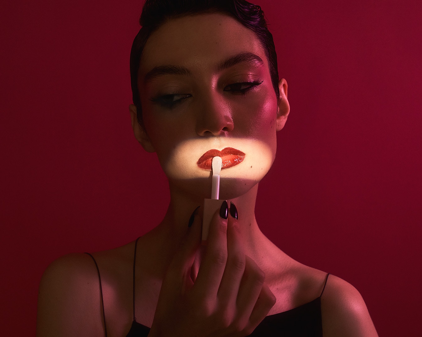 Primo piano di una donna che applica un lucidalabbra rosso brillante con un applicatore bianco, illuminata su uno sfondo rosso intenso, per un look audace e glamour.