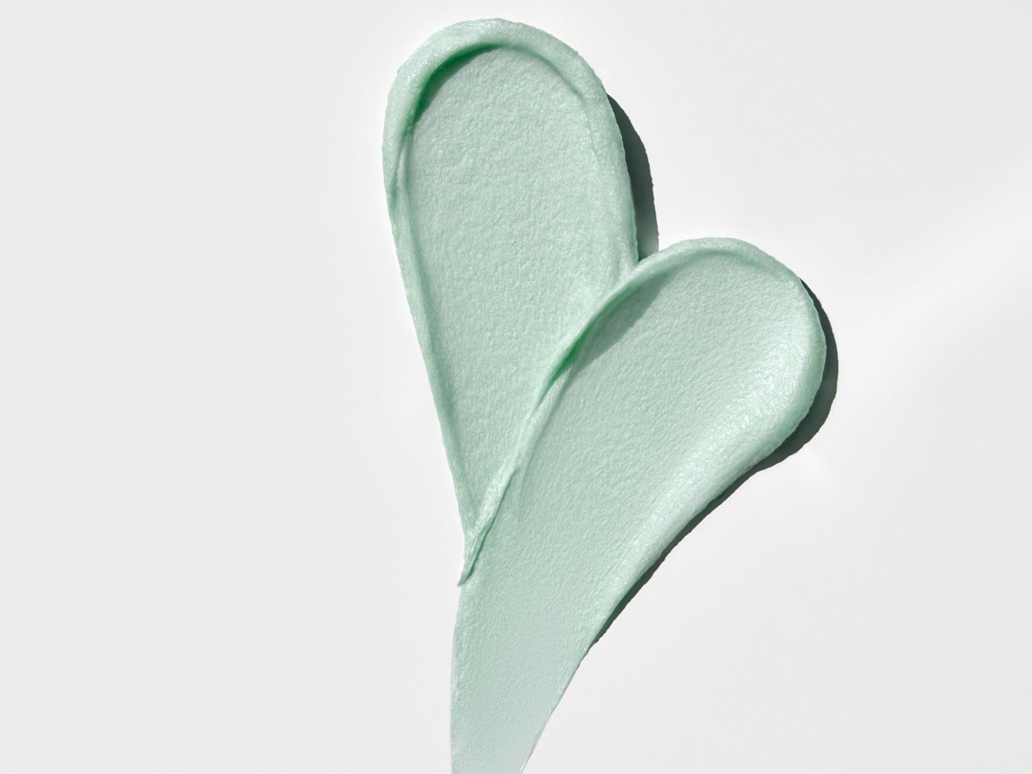 Nahaufnahme von zwei hellgrünen, herzförmigen Klecksen einer Creme oder Maske auf einem weißen Hintergrund.