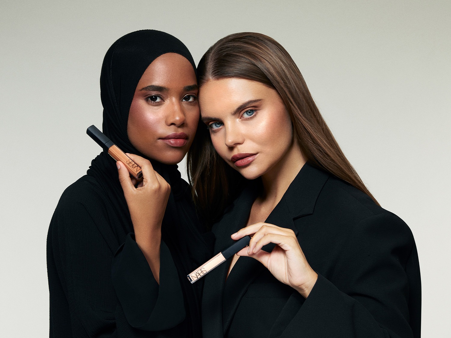 Zwei Models präsentieren NARS Concealer: Eine Frau mit Hijab hält einen beigen Concealer, während die andere einen helleren Farbton in der Hand hält.