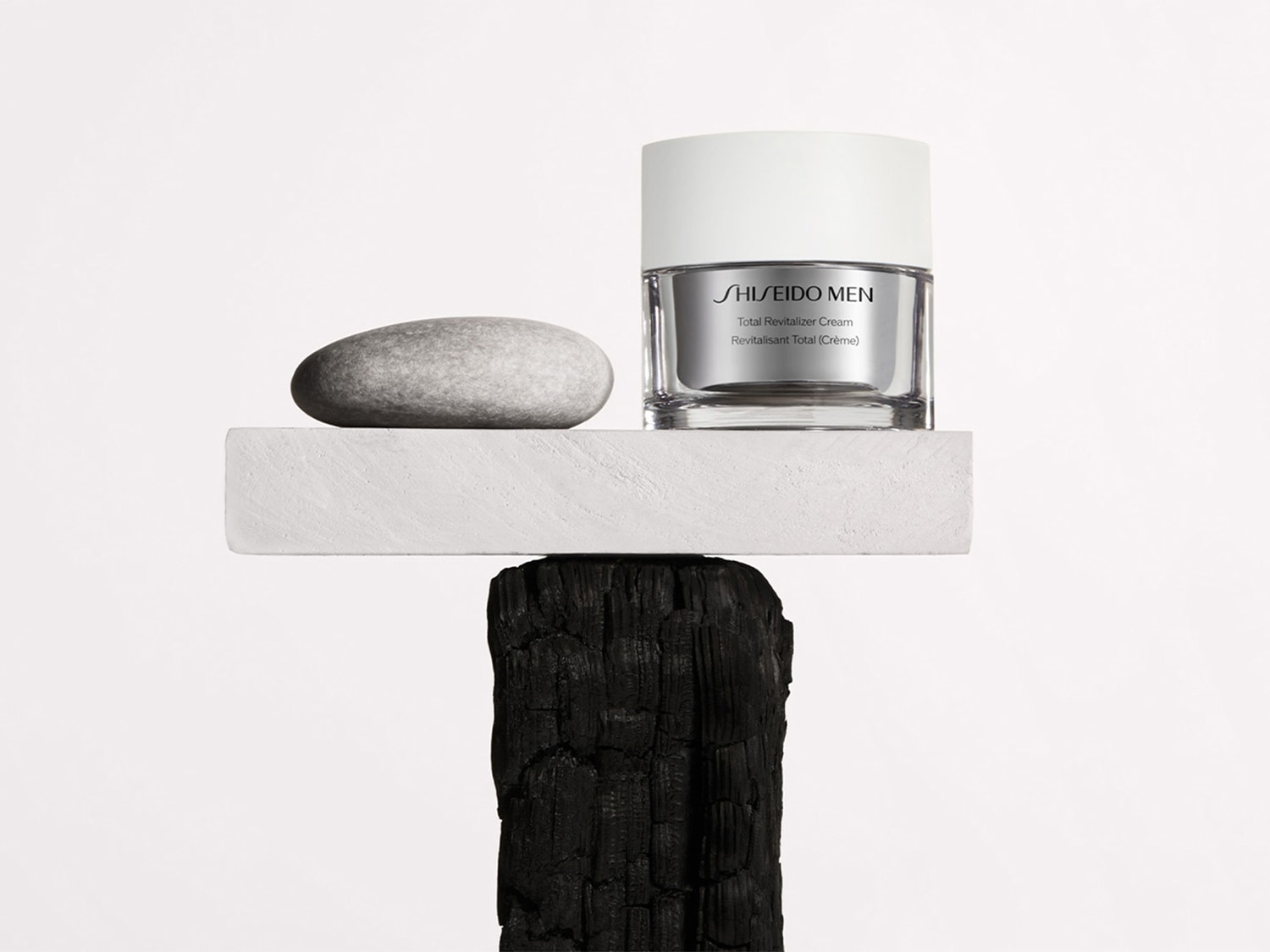 Shiseido Men Total Revitalizer Cream in einem durchsichtigen Tiegel mit silbernem Etikett und weißem Deckel, präsentiert auf einem weißen Sockel mit einem grauen Stein, der auf einem verkohlten Holzstück balanciert.