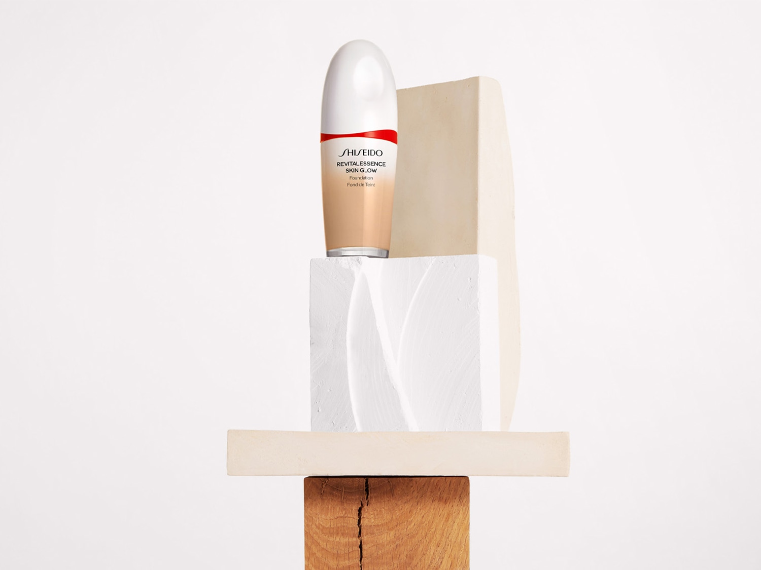 Shiseido Revitalessence Skin Glow Foundation in einer hellbraunen Flasche mit weißem Deckel und roter Aufschrift, die auf einem Stapel von geometrischen Formen aus Holz und Gips steht.