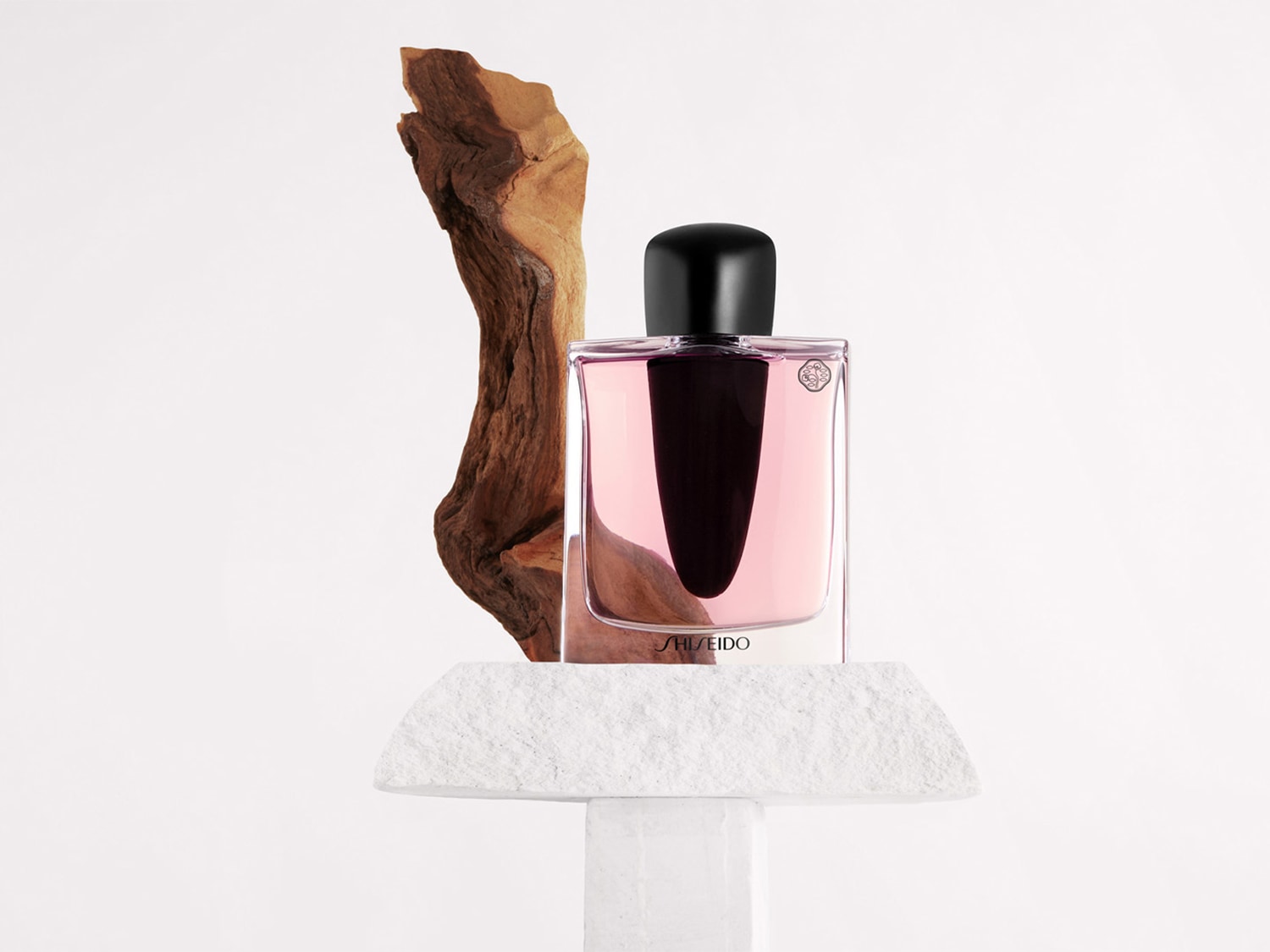 Ein rechteckiger, transparenter Shiseido-Parfümflakon mit rosa Flüssigkeit und schwarzem Verschluss steht auf einem weißen Steinsockel, begleitet von einem Stück Holz.