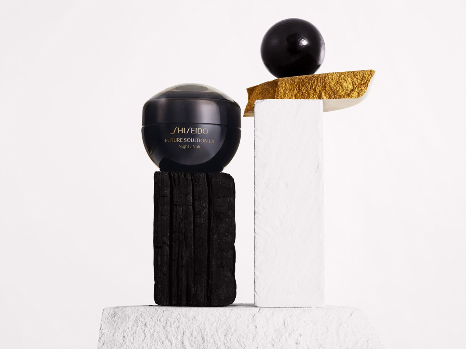 Schwarzer Tiegel Shiseido Future Solution LX Nachtcreme auf einem dunklen Holzsockel, darüber eine schwarze Kugel auf einem goldenen Akzent, der auf einem weißen Sockel ruht.