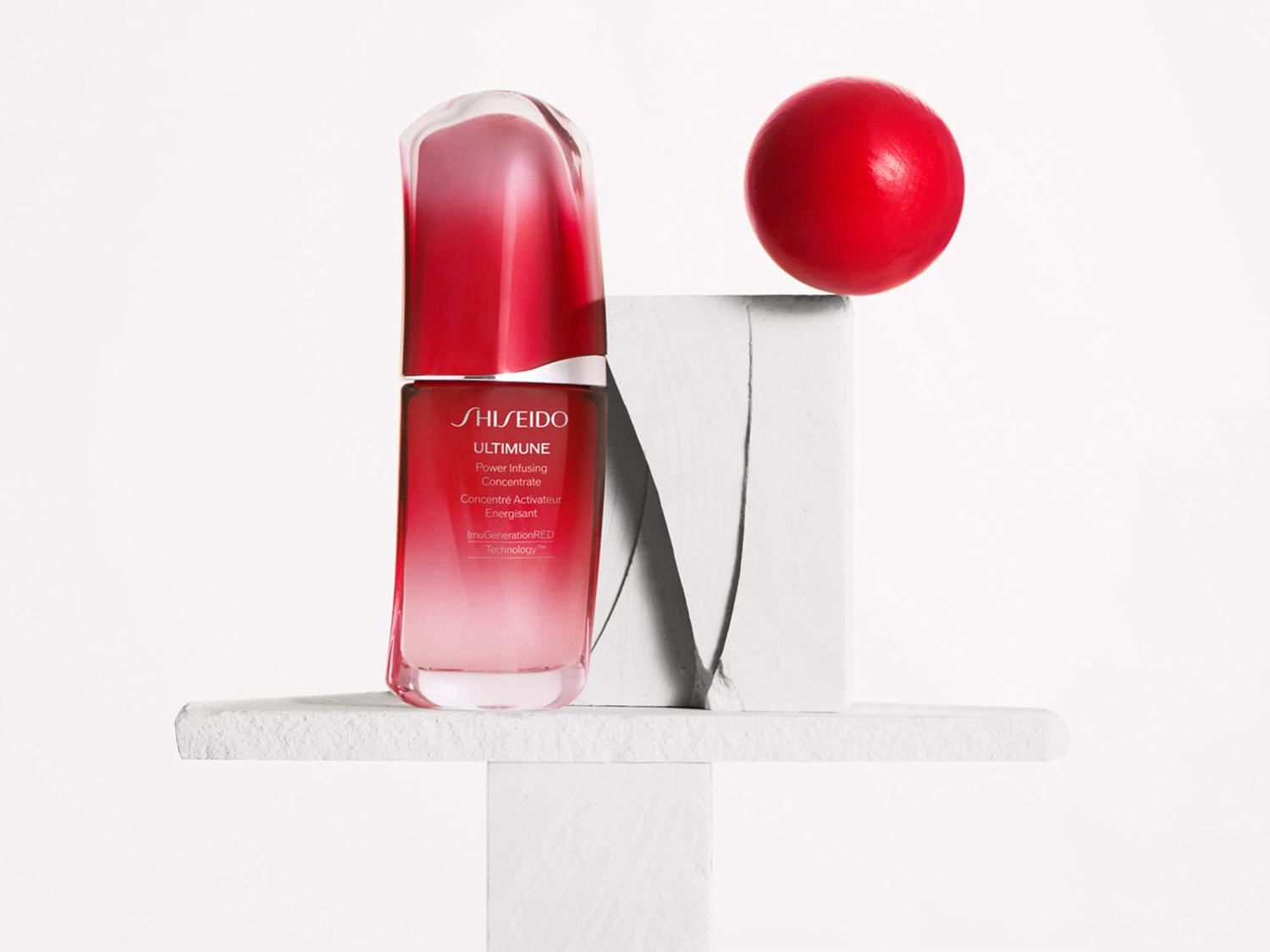 Shiseido Ultimune Power Infusing Concentrate in einer rot getönten Glasflasche, die auf einem weißen Sockel steht, mit einer roten Kugel im Hintergrund.